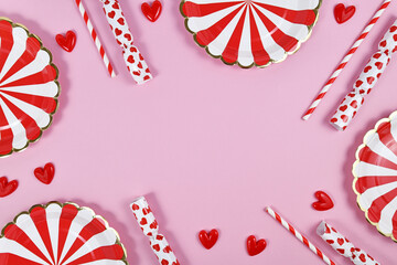Fototapeta premium Party border with red paper plates, heart ornaments, candy and drinking straws on pink background with copy space