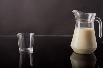 Glass jug with milk and empty glass cup. Glassware on reflective desk.