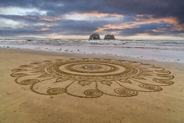 Intricate mandala star sand design on the ocean beach with Twin Rocks in the ocean and beautiful sky.