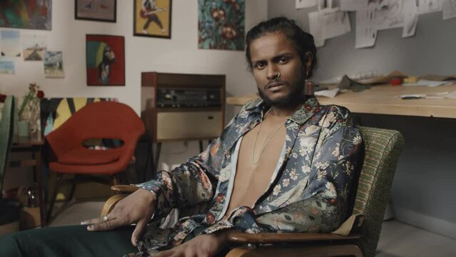 Portrait of handsome young Indian designer or artist in unbuttoned floral printed silk shirt with painted black nails sitting in armchair in creative messy workshop looking at camera
