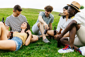 Group of multiracial friends hanging out and talking outdoors in public park - Diverse young people drinking beer together while relaxing outdoors in summer