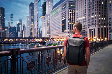Naklejka premium Rear view of man with backpack while walking on bridge and looking around. Illuminated city with skyscrapers at twilight. .
