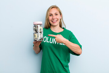young adult blonde woman smiling cheerfully, feeling happy and pointing to the side. volunteer and...