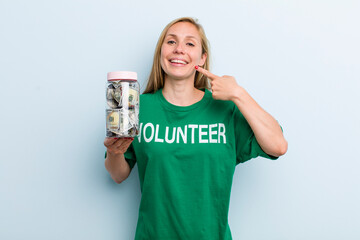 young adult blonde woman smiling confidently pointing to own broad smile. volunteer and donation...