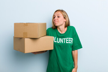 young adult blonde woman shrugging, feeling confused and uncertain. volunteer and donation concept