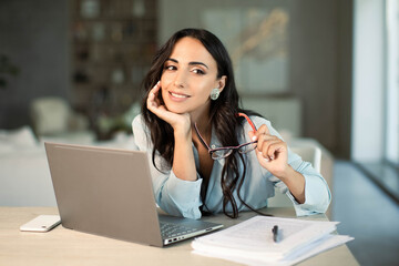 Smiling successful female using laptop computer indoors at home office, making notes on papers,...