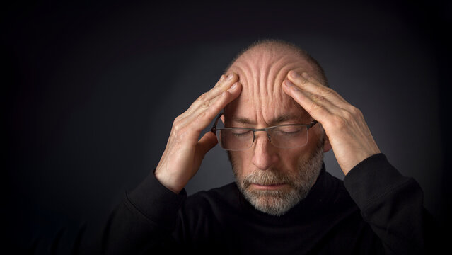 Tired - 60 Years Old  Man With A Beard And Glasses Massaging His Forehead -  A Headshot Against A Black Background With A Copy Space