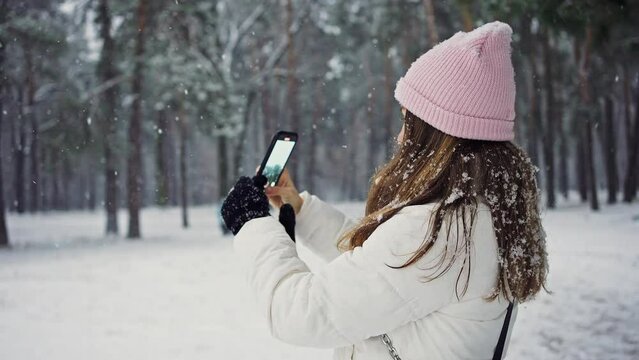 Young lady blogger making video on smartphone, recording snowfall nature during walk in winter forest, tracking shot