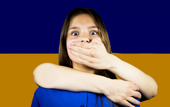 Sad little girl covered with Ukraine flag. Concept of standing with Ukrainian nation in war with Russia. Violence. A man's hand closes the child's mouth. - Powered by Adobe