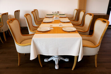 A restaurant consisting of brown wooden chairs and a table with a white fabric cover. Empty catering plates and forks, spoons, knives on a white tablecloth.	
