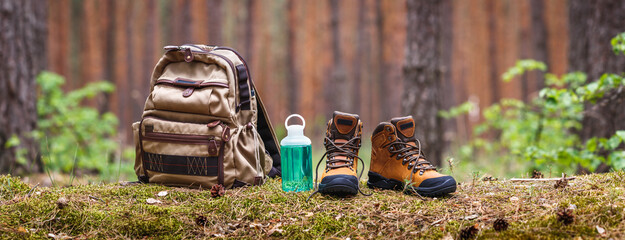 Hiking and camping equipment in forest. Backpack, water bottle and leather ankle boots. Panoramic...