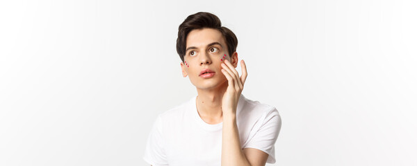 Fototapeta na wymiar People, lgbtq community and lifestyle concept. Beautiful young gay man applying glitter under eyes for pride party, standing over white background