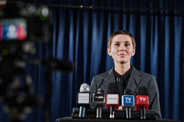 Young businesswoman with short hair standing at tribune with media microphones and performing...