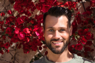 Portrait of handsome young man, with blue eyes and perfect smile, with beard and smiling green t-shirt with some red flowers in the background. Concept beauty, fashion, smile, perfect teeth, flowers.