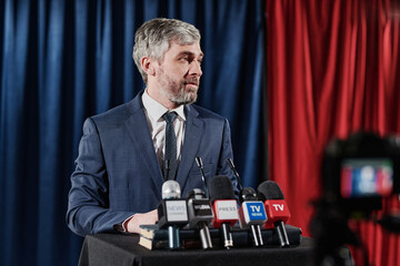 Mature bearded businessman standing at stage with microphones and giving an interview during online...