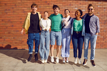 Happy diverse friends having fun and sharing positive emotions during city walk in spring or summer. Group of joyful young people in jeans jumping all together on street with brick wall in background