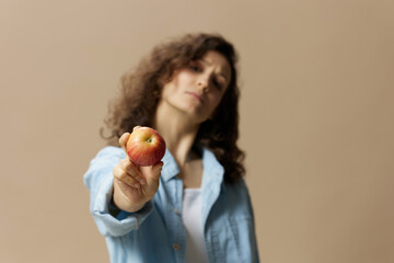 Cute cheerful happy curly beautiful female in jeans casual shirt pulls apple at camera posing isolated on over beige pastel background. Healthy food. Natural eco-friendly products concept. Copy space