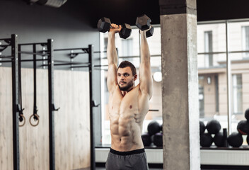 Fototapeta na wymiar Athletic man with a naked torso trains his shoulders with dumbbells in his hands. Workout in the gym. Bodybuilding and fitness, healthy lifestyle