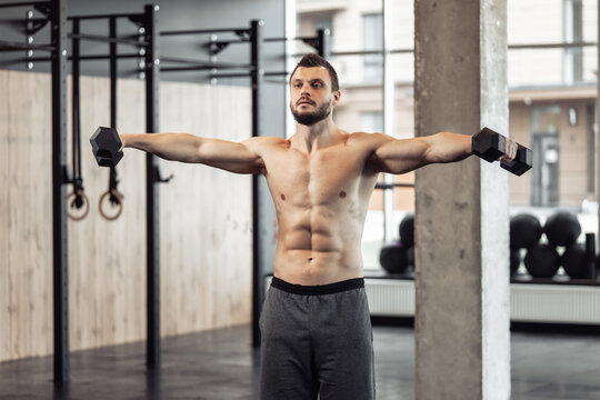 Athletic man with a naked torso trains his shoulders with dumbbells in his hands. Workout in the gym. Bodybuilding and fitness, healthy lifestyle
