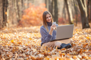 Young female freelancer or student with a laptop and smartphone sits on fallen leaves in autumn forest. Remote work or education