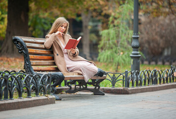 Cute beautiful woman reading book, sitting on bench in autumn park
