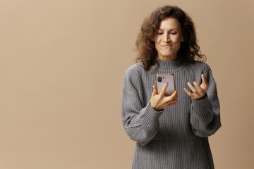 Evil irritated curly beautiful female in gray casual sweater squeeze phone with fury posing isolated on over beige pastel background. Social media, network, distance communication concept. Copy space