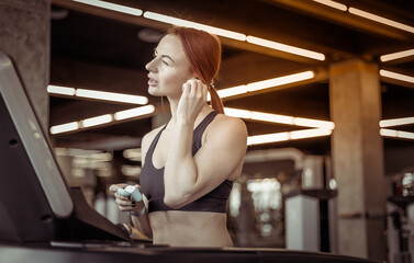 Obraz na płótnie Canvas Portrait of a red-haired fitness woman with earphones in a modern gym. Fit slim woman works out on a treadmill, cardio workout, healthy lifestyle.
