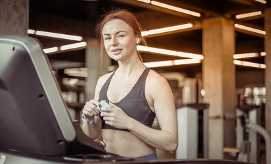 Portrait of a red-haired fitness woman with earphones in a modern gym. Fit slim woman works out on a treadmill, cardio workout, healthy lifestyle.