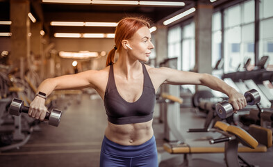 Fototapeta premium Attractive red-haired fitness woman trains shoulder muscles with dumbbells in a modern gym. Bodybuilding and Fitness