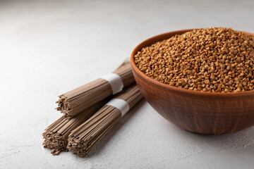 Buckwheat porridge in a bowl and buckwheat soba noodles on a white marble background. Raw buckwheat. Ingredients for gluten free cereal. Healthy food. Place to copy.
