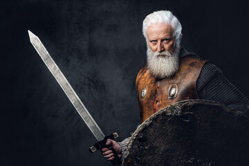 Portrait of gray haired medieval knight with long beard dressed in chain mail holding sword and...
