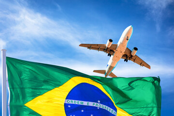 Brazil flag blowing in the wind and plane landing with blue sky and clouds in the background.