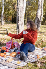 Beautiful woman in red sweater on a picnic in a autumn forest using mobile