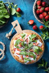 Summer season. Pizza with cheese strachatella, strawberries, arugula and mint with spices on a bright turquoise background. Rustic. Top view. Background image, copy space
