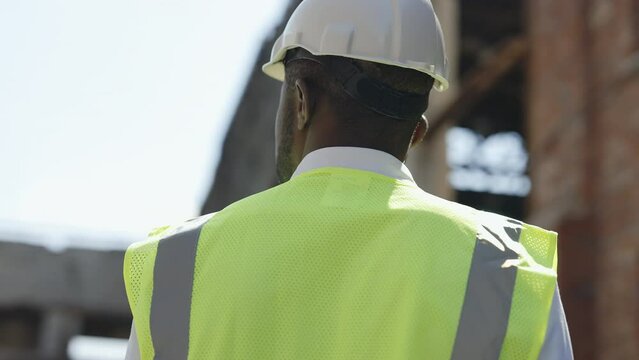 Back view of focused african american architect in workwear and helmet looking around while standing at construction site. Concept of engineering, development and business.