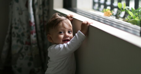 Baby next to home window looking outside. Infant boy leaning window learning to stand