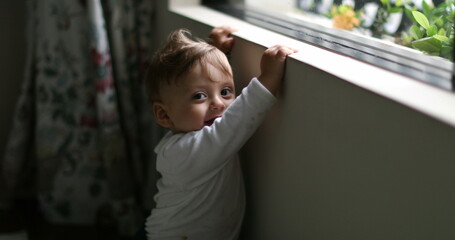 Baby next to home window looking outside. Infant boy leaning window learning to stand