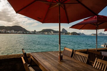 Rio de Janeiro, Brazil. Beach and sea and buildings on the edge of Copacabana Bay, mountains and Sugar Loaf Mountain seen from Copacabana Fort. Wooden tables and chairs with umbrellas.