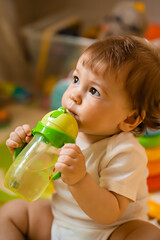 Cute little boy drinking water from a training bottle with a straw, home interior, lifestyle, vertical photo