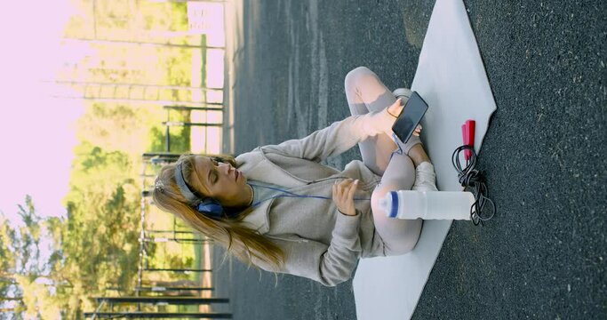 Young Woman Meditating After Outdoor Workout. She Sits In Headphones And Listens To Relaxing Music.