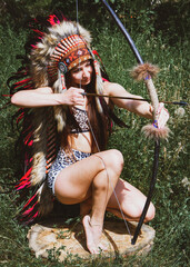 A sexy lady in an Indian roach shoots an arrow from a bow on a background of grass.