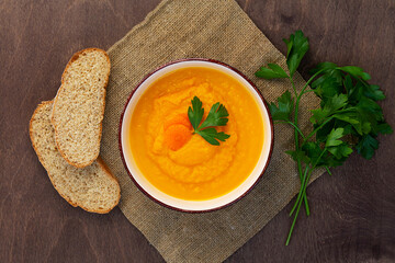 Carrot soup with fresh parsley with sackcloth on brown table. Bowl of tasty carrot cream soup on wooden background. Top view. Spring vegetable soup. Homemade pumpkin cream soup with bread.