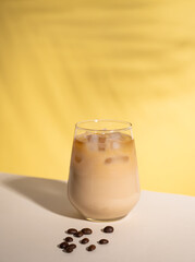 Iced coffee with milk in a glass with ice cubes and grains on a yellow background with palm leaf shadows. The concept of a cold summer drink. Copy space