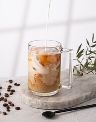 Iced coffee in a glass with pour cream, ice cubes and grains on a light marble background with morning shadows. The concept of a cold summer drink