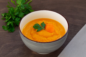 Grey bowl of tasty carrot cream soup with fresh carrots and herbs on wooden background. Carrot soup with parsley on brown table. Spring vegetable soup. Homemade cream soup with copy space.