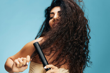 Joyful happy Latin curly woman using hair straightener, looking aside, posing isolated on blue wall...