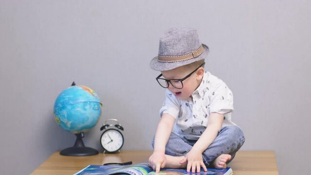 happy child reading book sitting on wooden table with earth globe, alarm clock on grey background. funny smart and intelligent caucasian kid boy in hat and eyeglasses pointing finger in blue picture