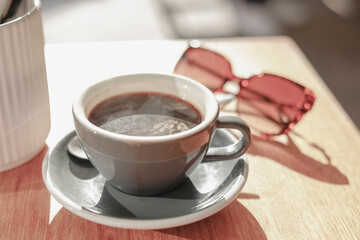 A cup of coffee sitting on a table with sunglasses in the background at a cafe