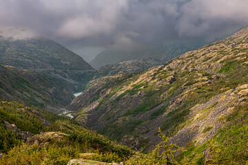 Naklejka premium Skagway, Alaska, USA - July 20, 2011: Klondike highway to Canada. Down the road, the valley with small pond under heavy cloudscape over green mountain flanks.