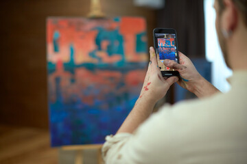 Young modern artist holding phone taking pictures of his abstract painting for social network blog. Close up rear view